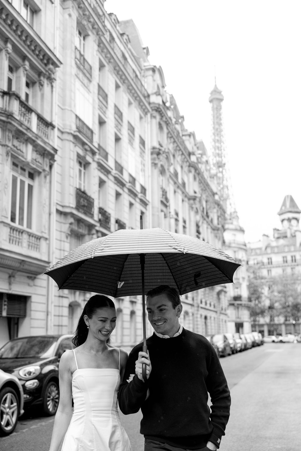 A couple walks down a Parisian street with umbrellas as the Eiffel Tower. Rainy paris photoshoot. Rainy cinematic paris photos. film photo