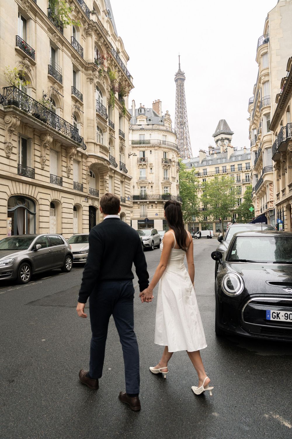 A couple walking on a Parisian street with the Eiffel Tower visible in the background through historic buildings.