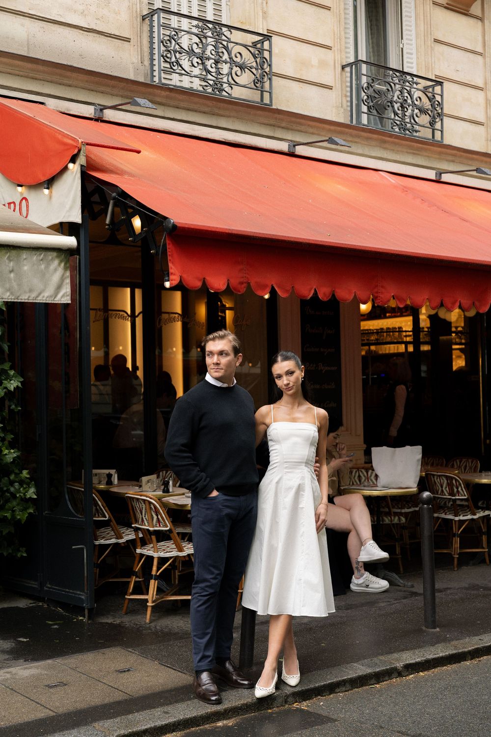 A couple poses for wedding photos outside a Parisian cafe with red awnings and classic bistro chairs.