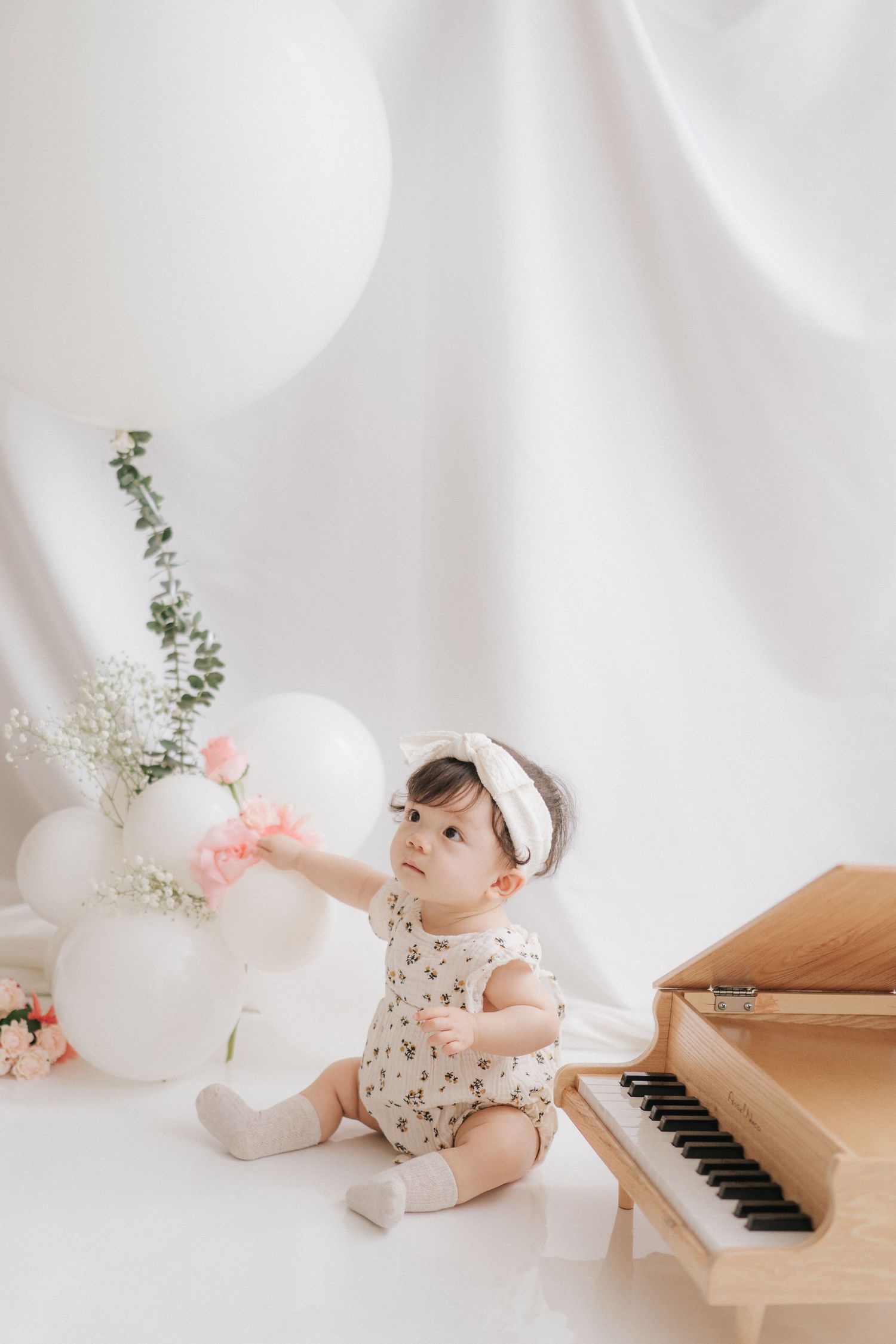 Baby in floral outfit plays near piano with white balloons and delicate floral arrangements in dreamy studio setting.