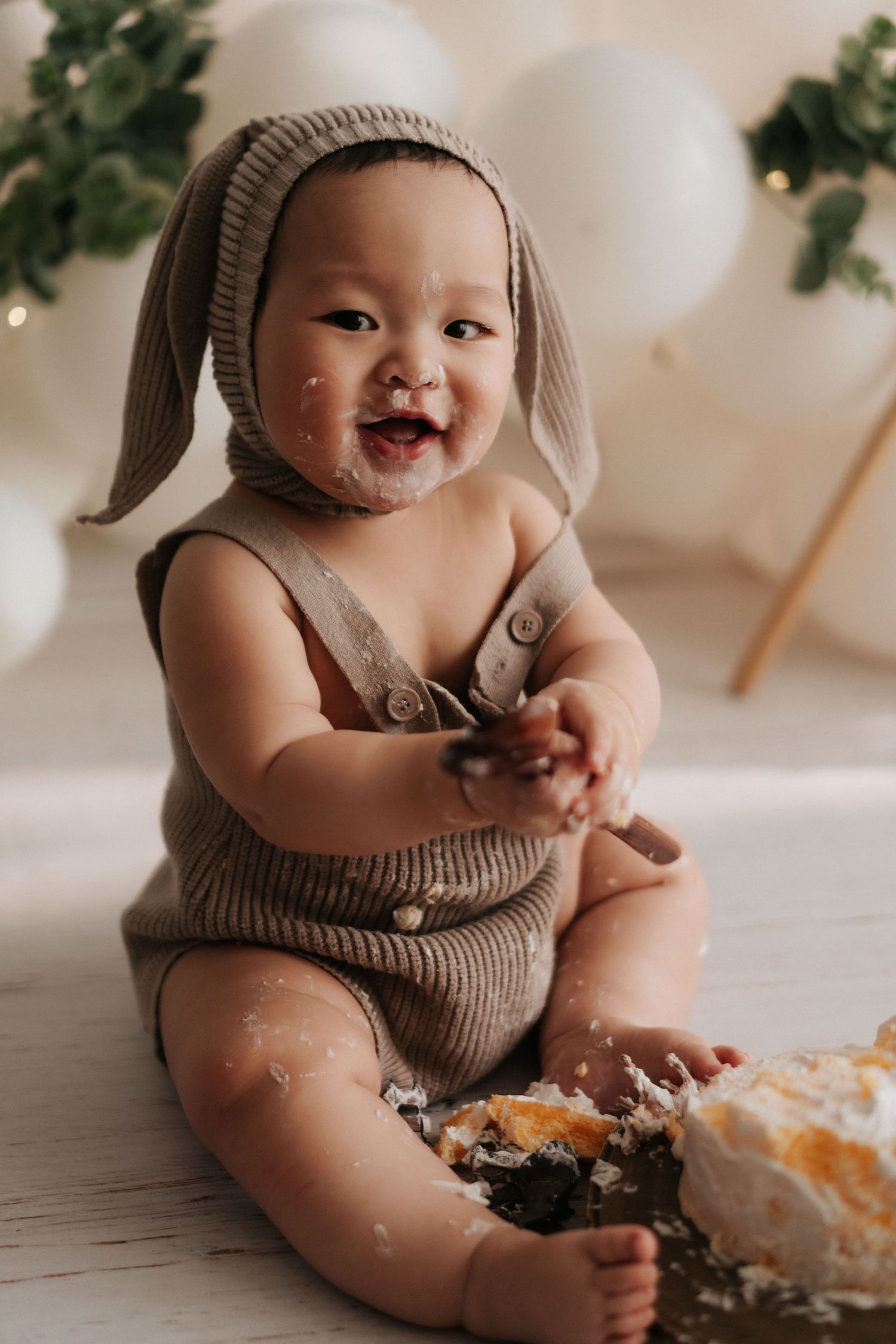 Happy baby wearing knitted bunny hat and romper sits on floor with smashed cake during first birthday photo shoot.
