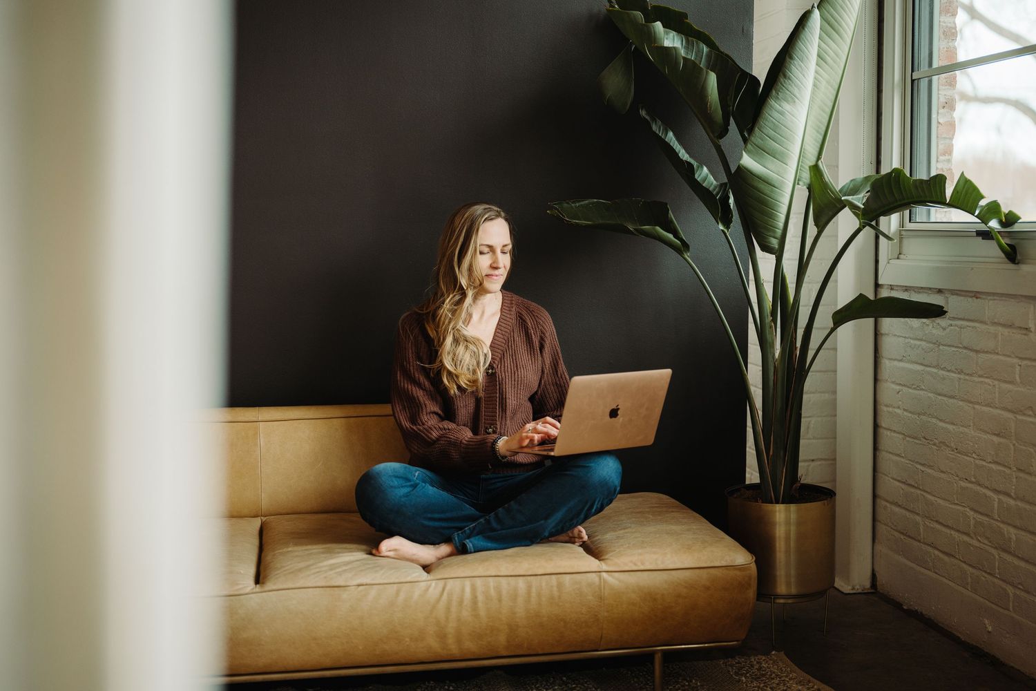 Person working on laptop while relaxing on comfortable beige couch near window with natural light and plants.