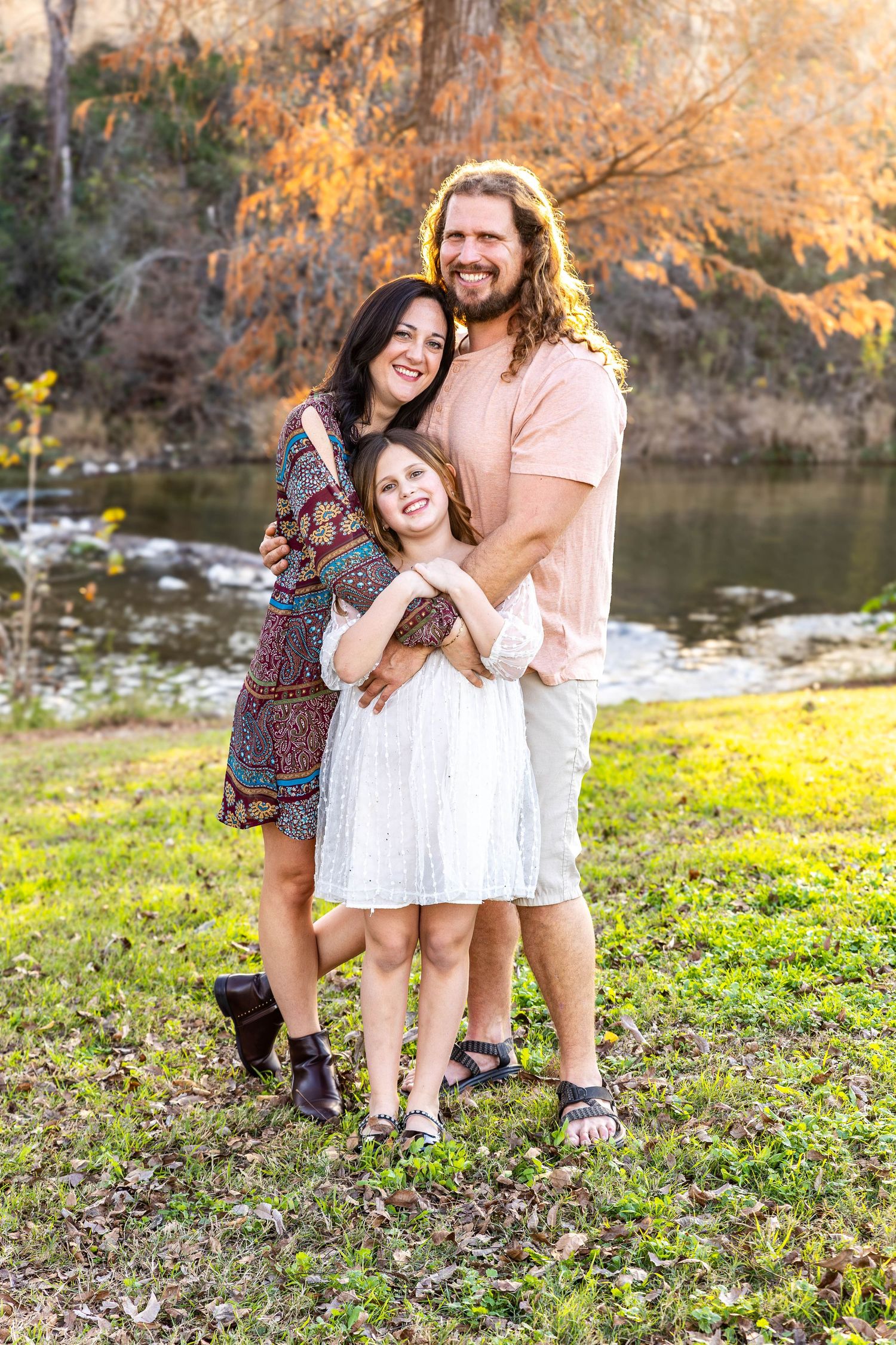 Family of three posing together by a lake during golden hour with autumn trees in background.
