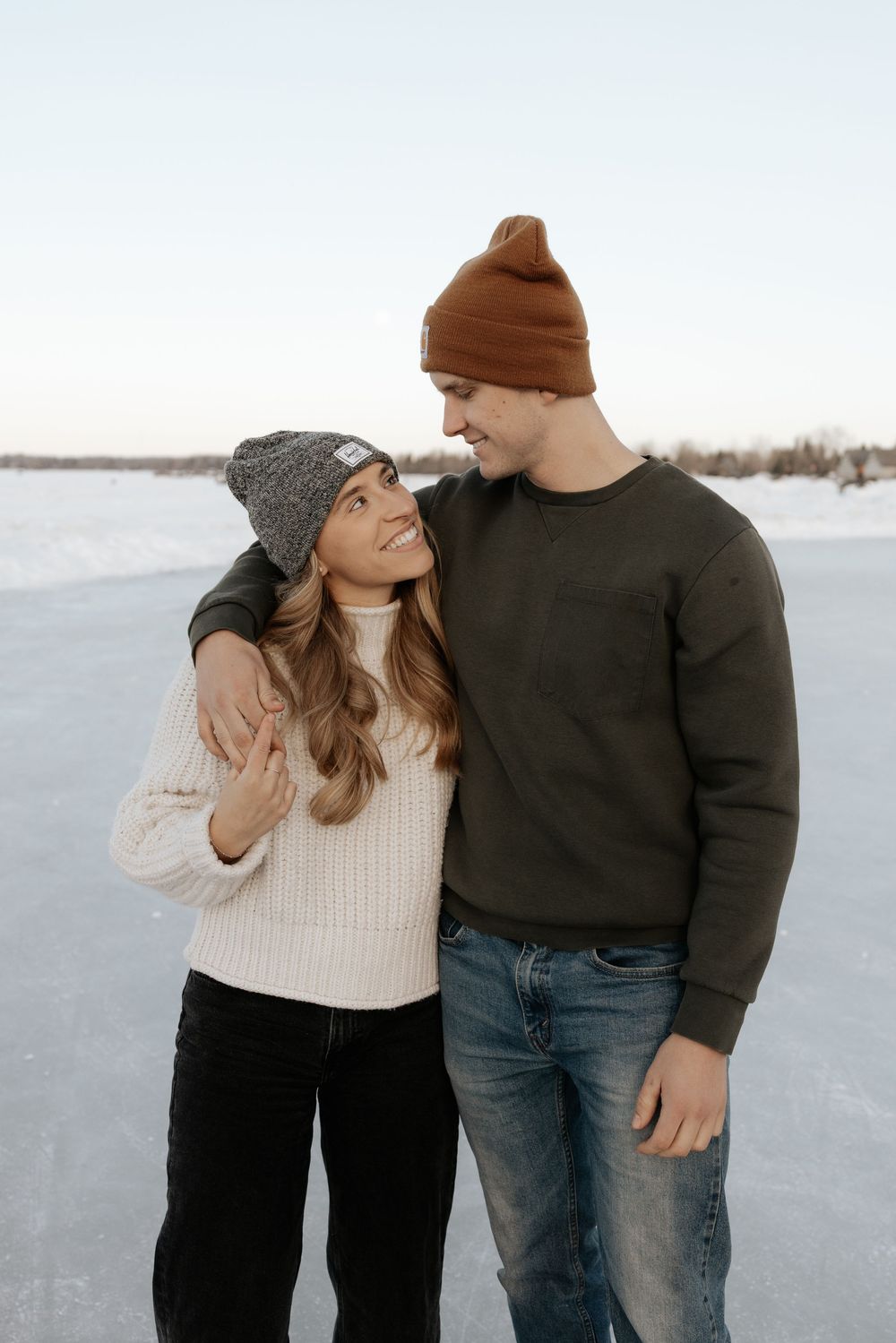 A couple in winter clothing embraces while standing on a snowy landscape in a romantic outdoor photoshoot.