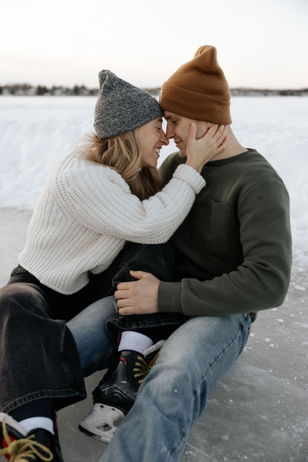 A couple sits close together on a snowy winter day wearing cozy knit hats and warm clothing.