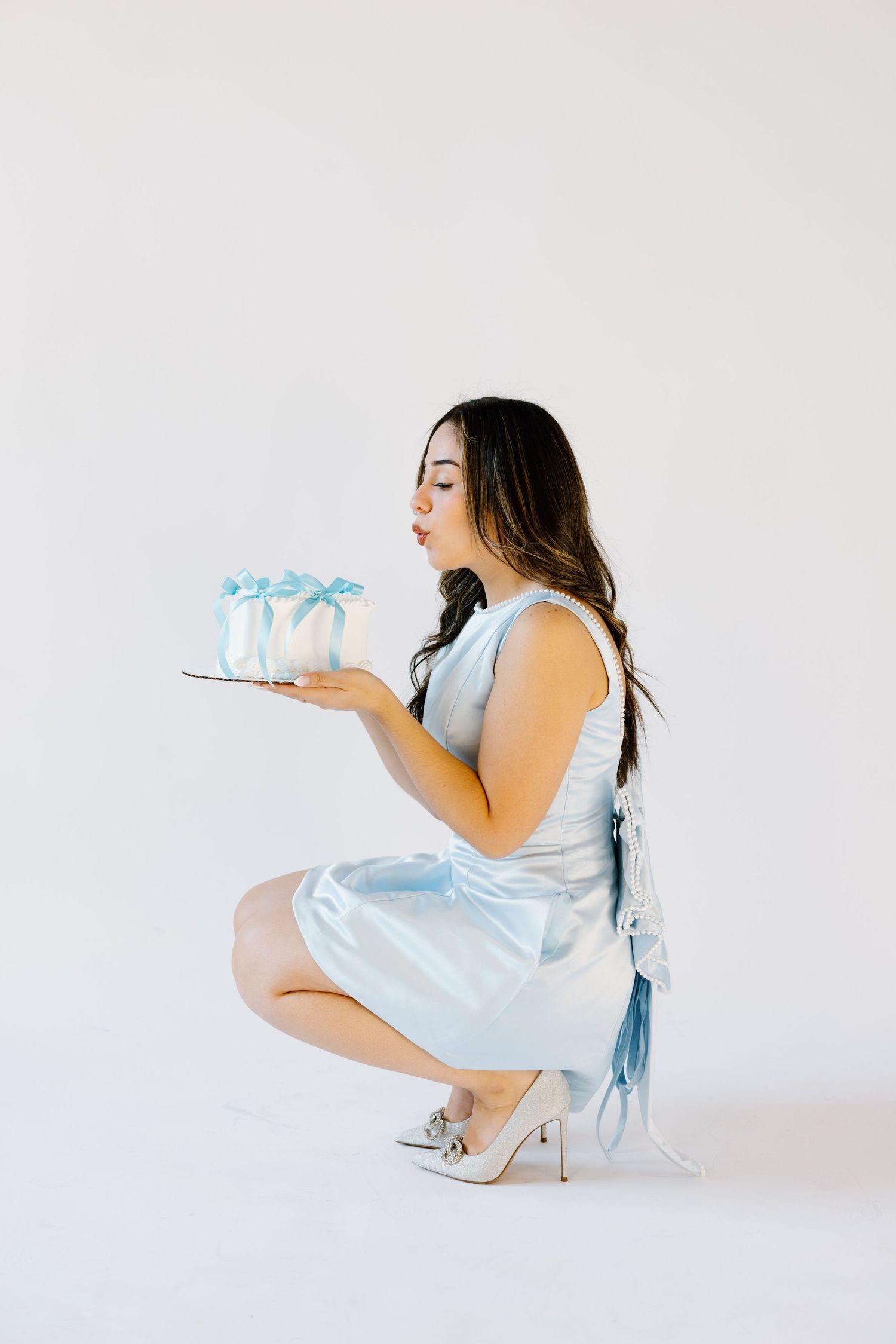 Woman in light blue dress crouches while holding decorated cake against white background.