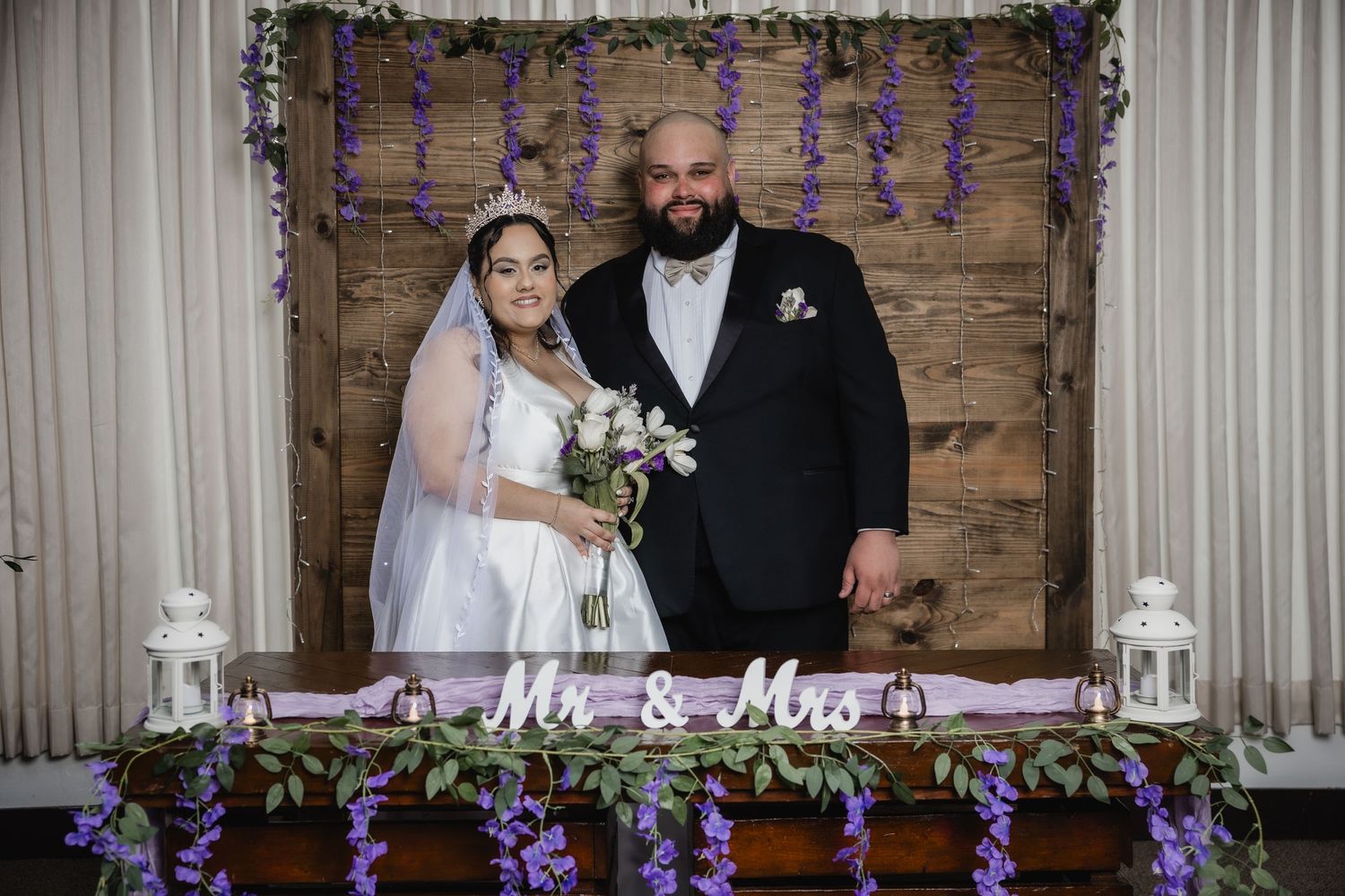 A wedding ceremony setup with rustic wood backdrop, purple floral decor, lanterns and Mr & Mrs sign on display table.