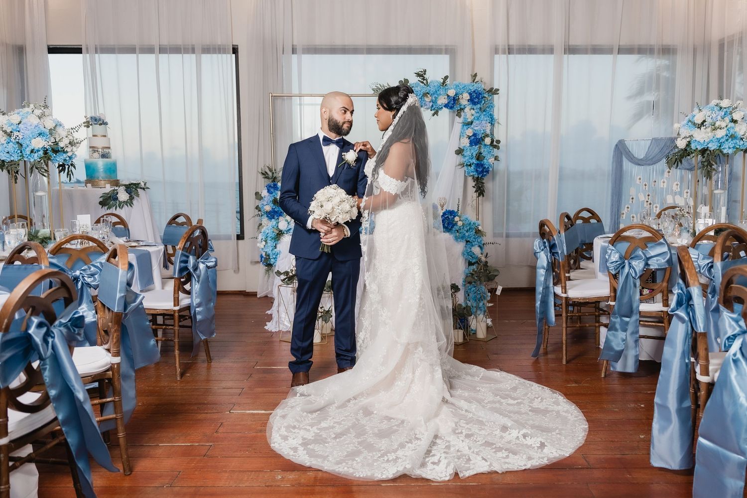 A couple shares their wedding ceremony moment under a blue floral arch in a rustic indoor venue with wooden floors.