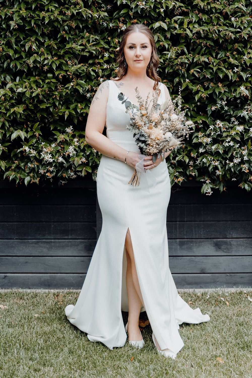Bride in elegant wedding gown with front slit and dried floral bouquet, captured by Apertured Media Auckland Wedding Photographer.