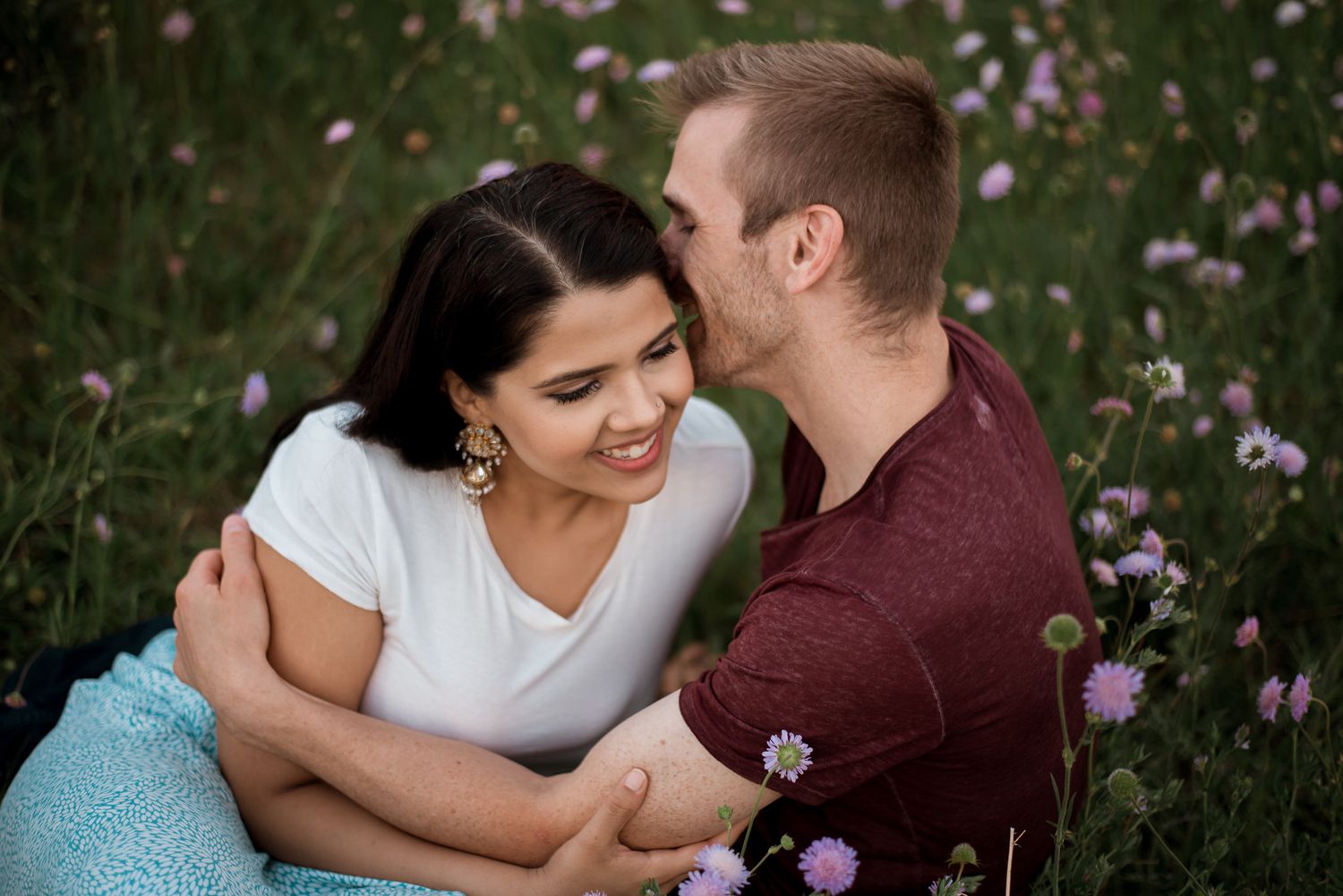 A couple shares an intimate moment while sitting together in a wildflower meadow on a sunny day.