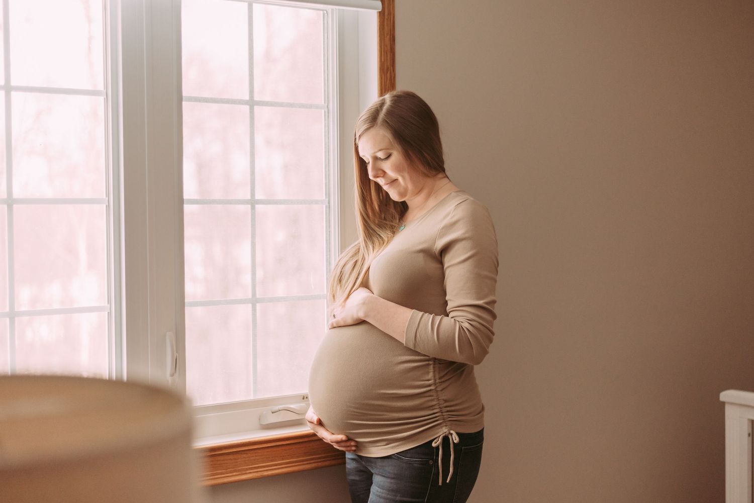 Expectant mother in beige sweater stands by window, holding her pregnant belly in a tender moment of contemplation.