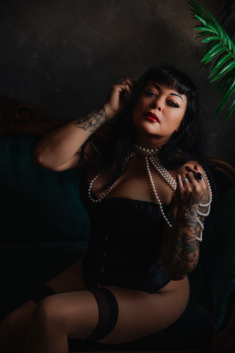 A Woman in a black corset and thigh highs poses dramatically with pearl necklaces in a dark and moody Utah boudoir photography session