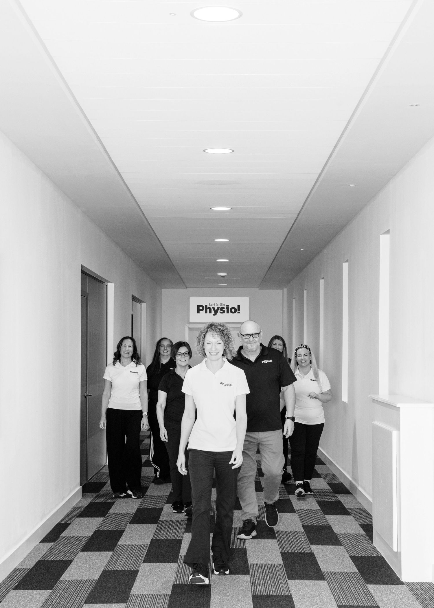 A group of colleagues stand together in a modern office hallway with checkered carpet and recessed lighting.