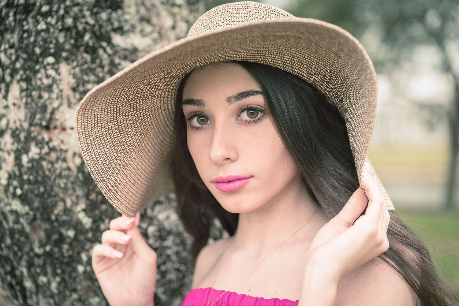 Portrait in soft lighting with straw hat and pink lipstick against a blurred tree background.