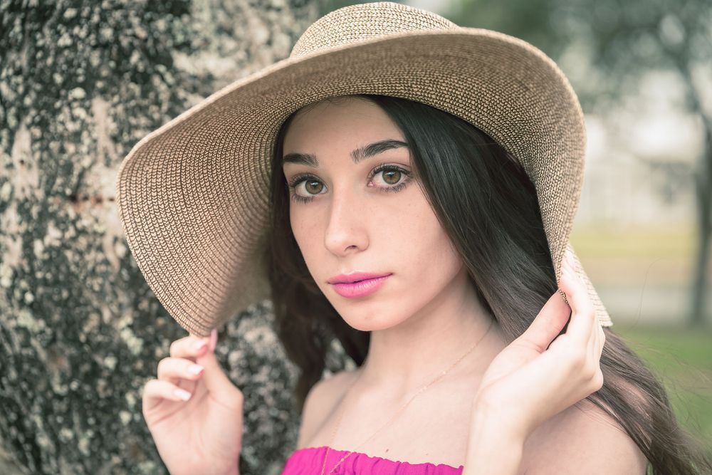 Portrait in soft lighting with straw hat and pink lipstick against a blurred tree background.