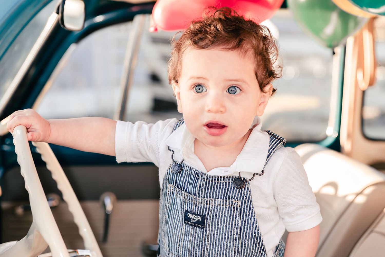 Toddler in striped overalls sitting in car seat with surprised expression.
