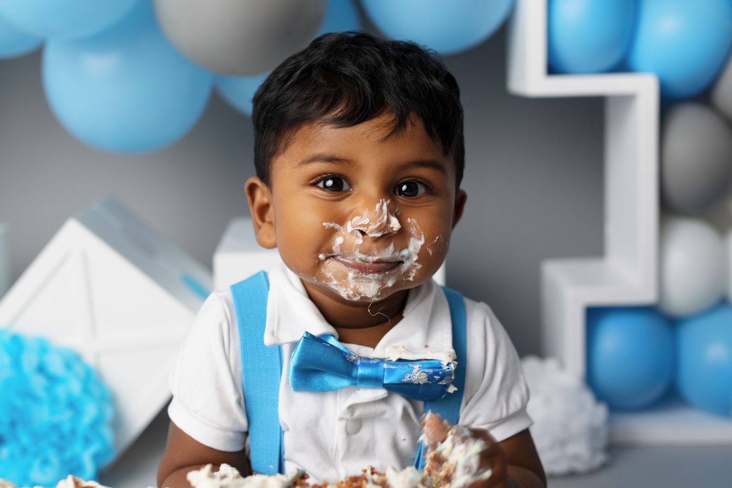 Young child with cake frosting on face at birthday party with blue balloons.