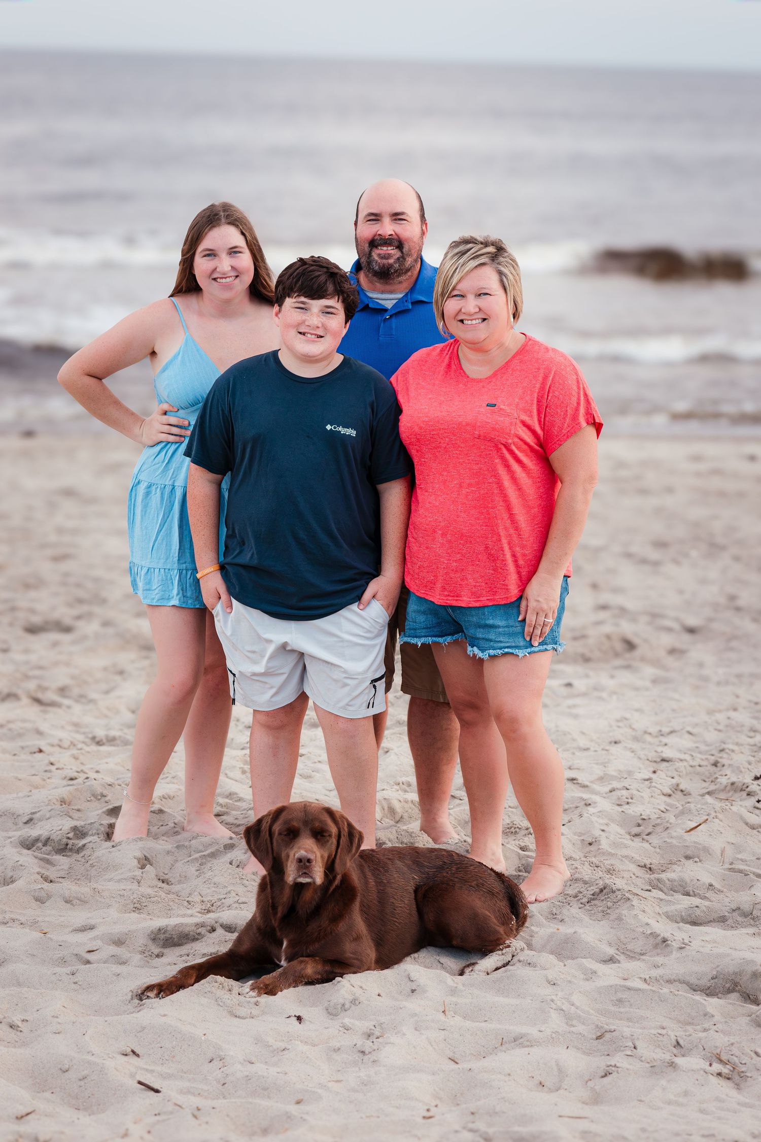 Beach family portrait with chocolate Labrador retriever resting in sand by the ocean.