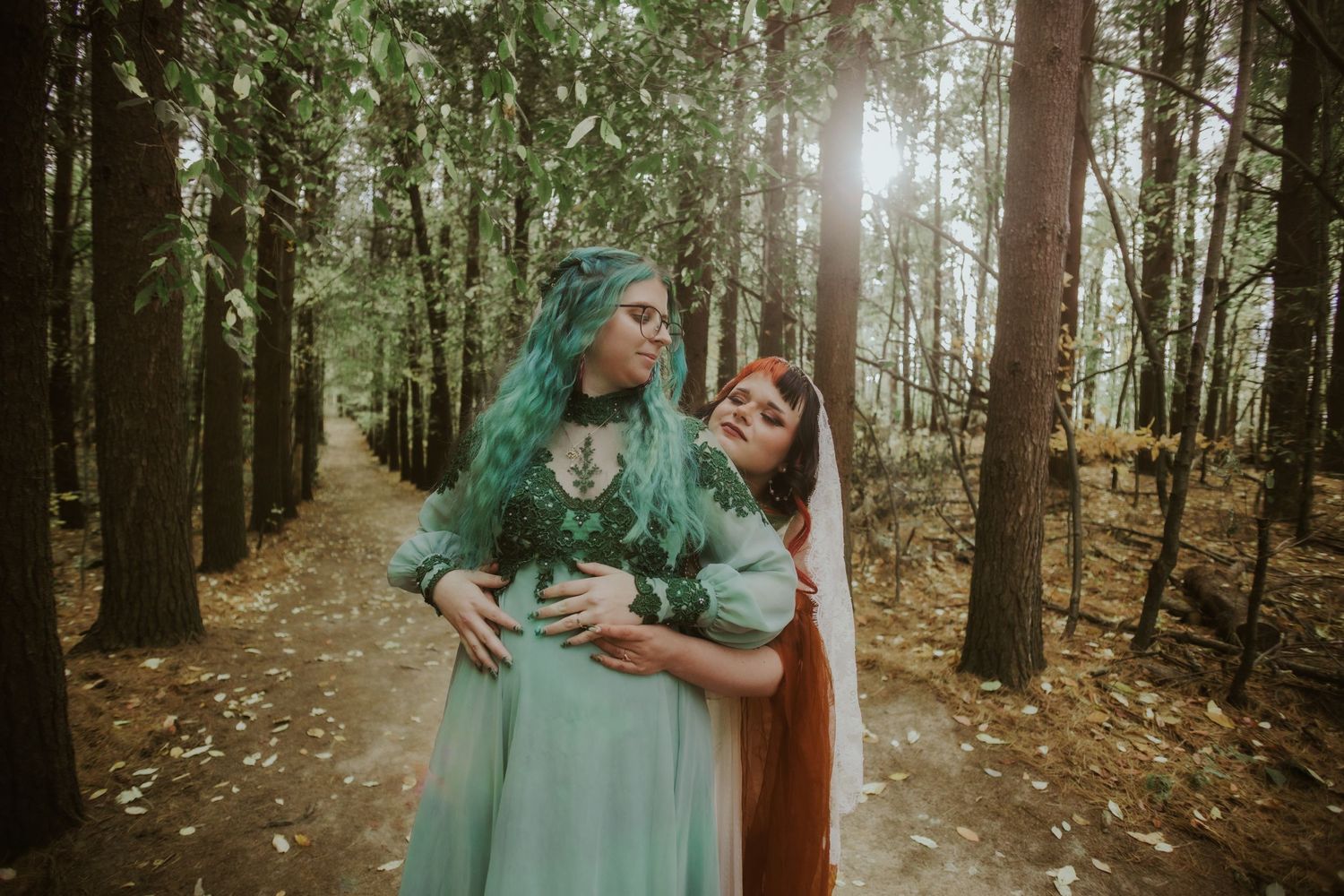 Two women in ethereal teal and white dresses pose playfully in a misty forest.