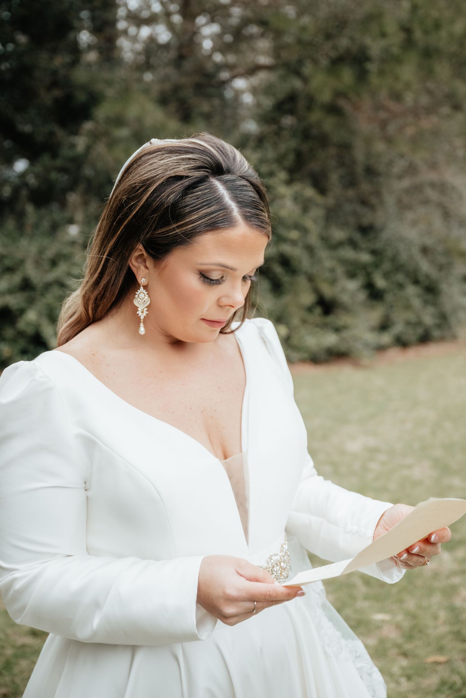 Elegant bride in white long sleeve gown with deep V-neck reading heartfelt letter outdoors before wedding ceremony.