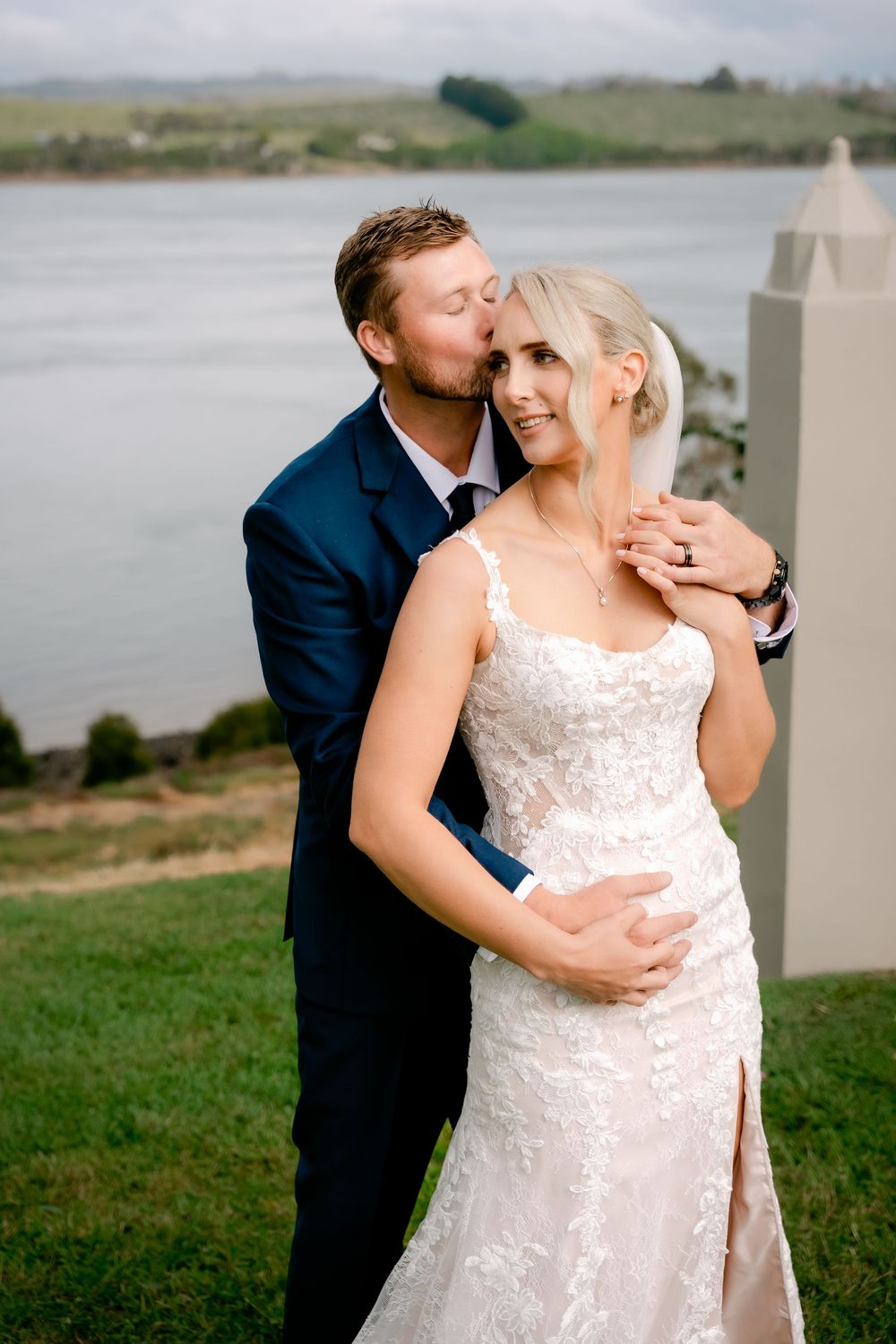 A romantic wedding couple embraces on a grassy hillside overlooking a serene waterfront on their special day.