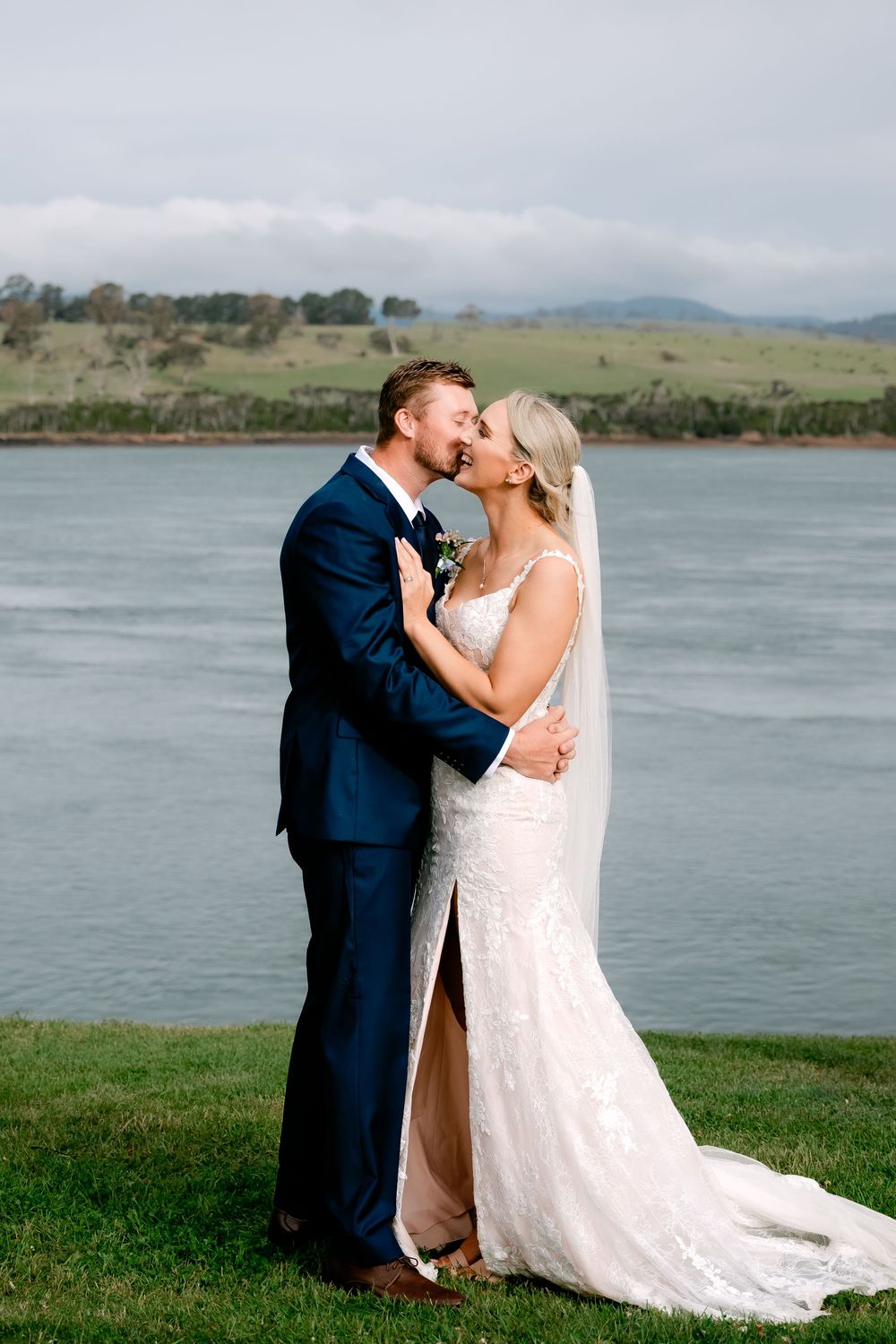 Newlyweds share a romantic kiss by a scenic lake with rolling hills in the background.
