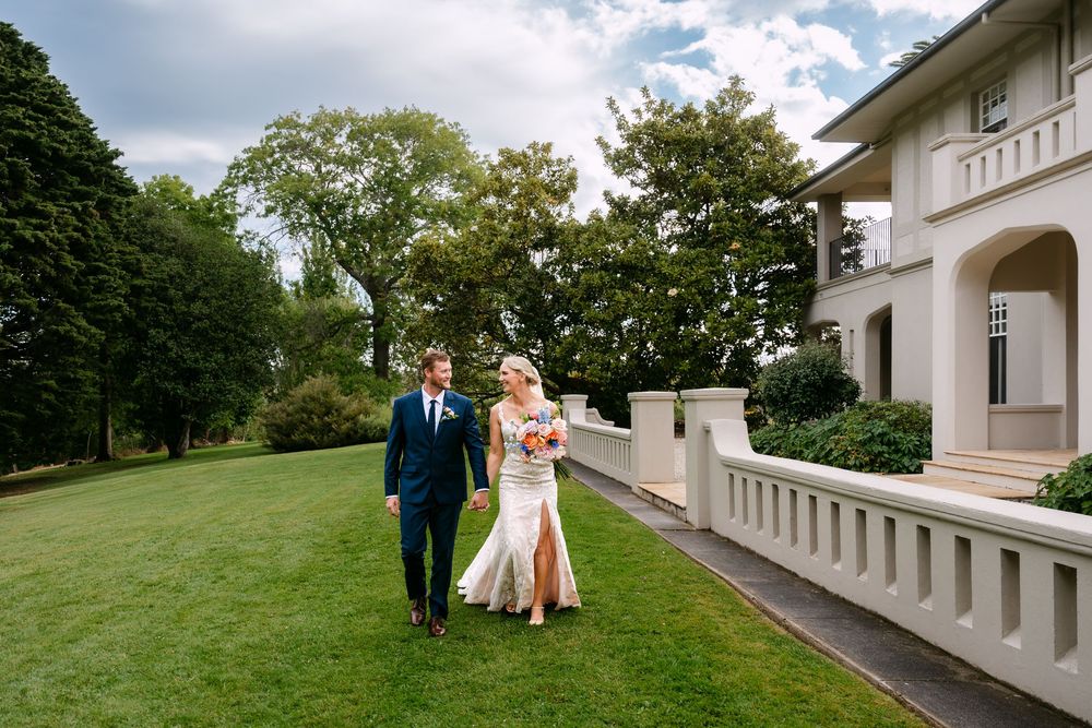 Elegant wedding couple poses on a grand white balcony with lush green landscaping in the background.