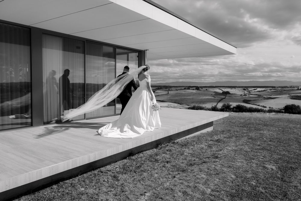 A black and white wedding photograph showing figures on a modern architectural terrace overlooking a dramatic landscape.
