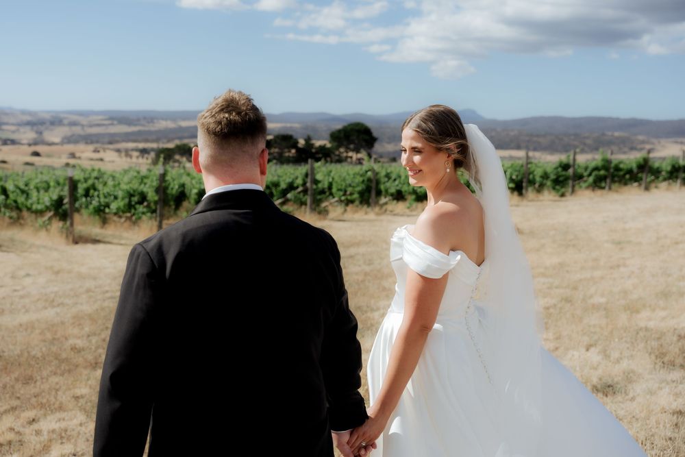A couple in wedding attire stands together on a hilltop overlooking vineyards and mountains on their special day.