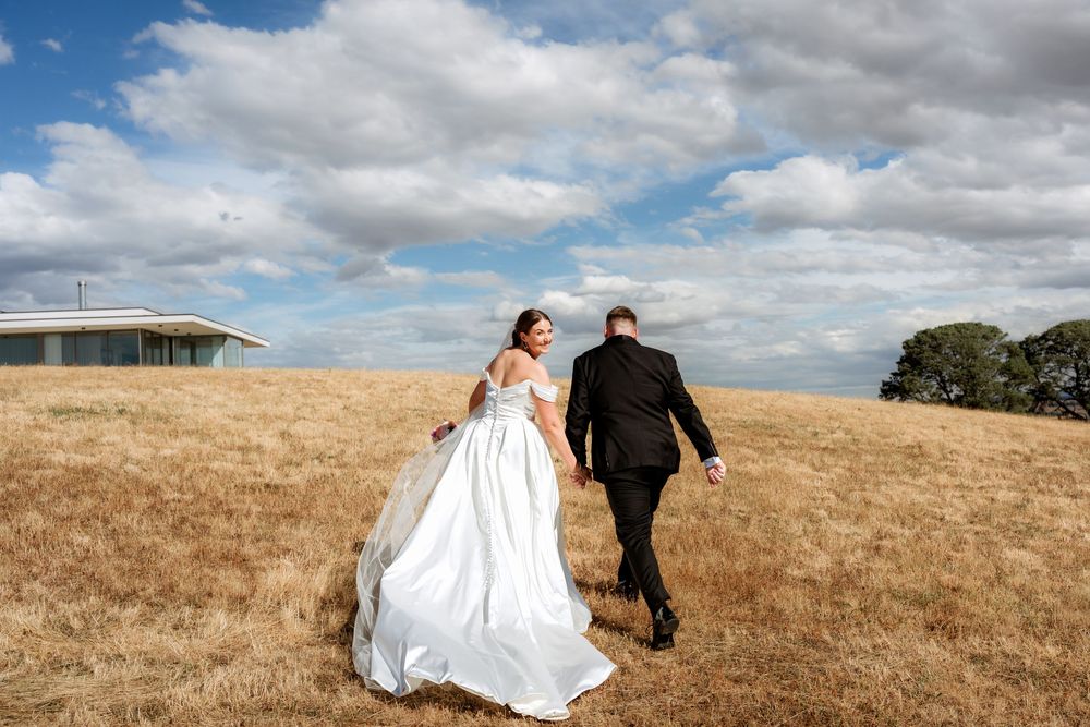 A couple in wedding attire walks hand in hand across a golden hillside under dramatic cloudy skies.