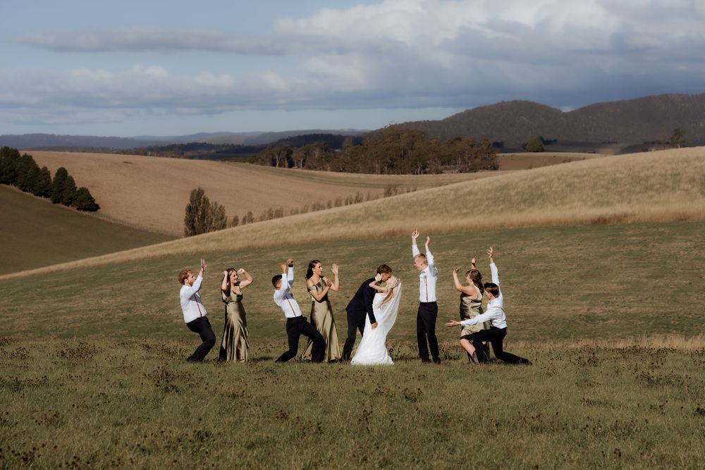 Wedding party poses playfully in a scenic mountain meadow with rolling hills and dramatic clouds in the background.