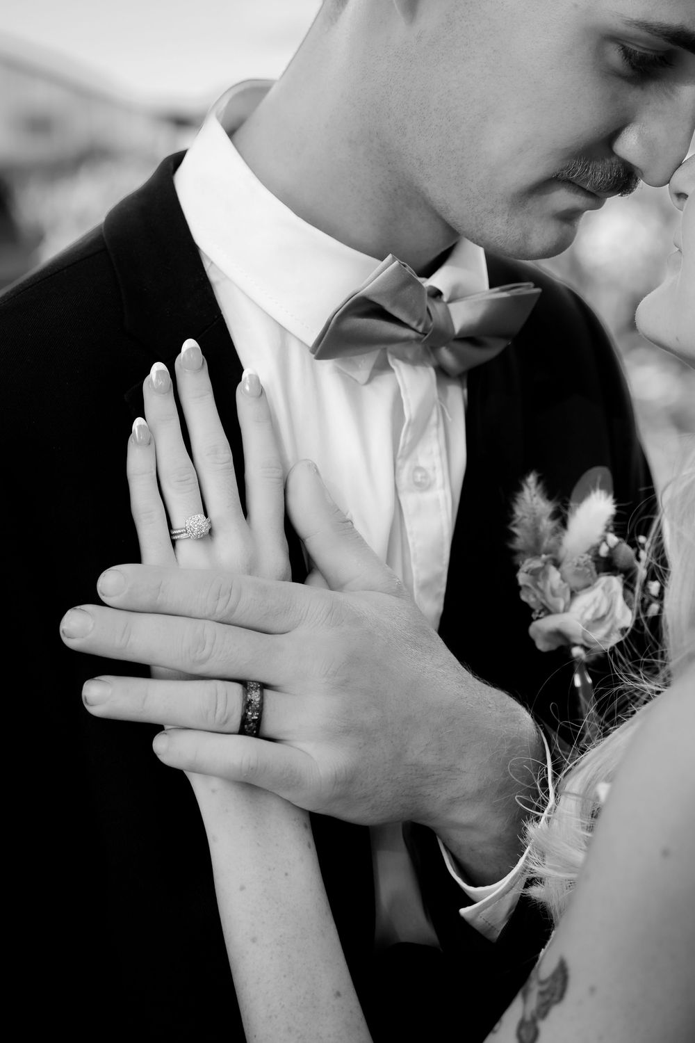 A black and white wedding photo showing hands with wedding rings and a boutonniere on the groom's lapel.