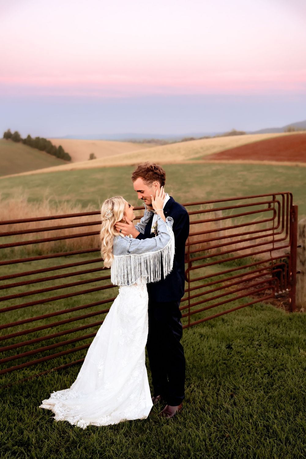 A couple in wedding attire embrace on a hilltop at sunset with rolling farmland in the background.