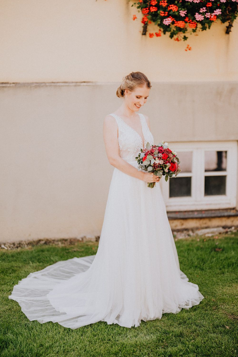 An elegant bride in a flowing white gown holds a red and pink bouquet against a cream wall.