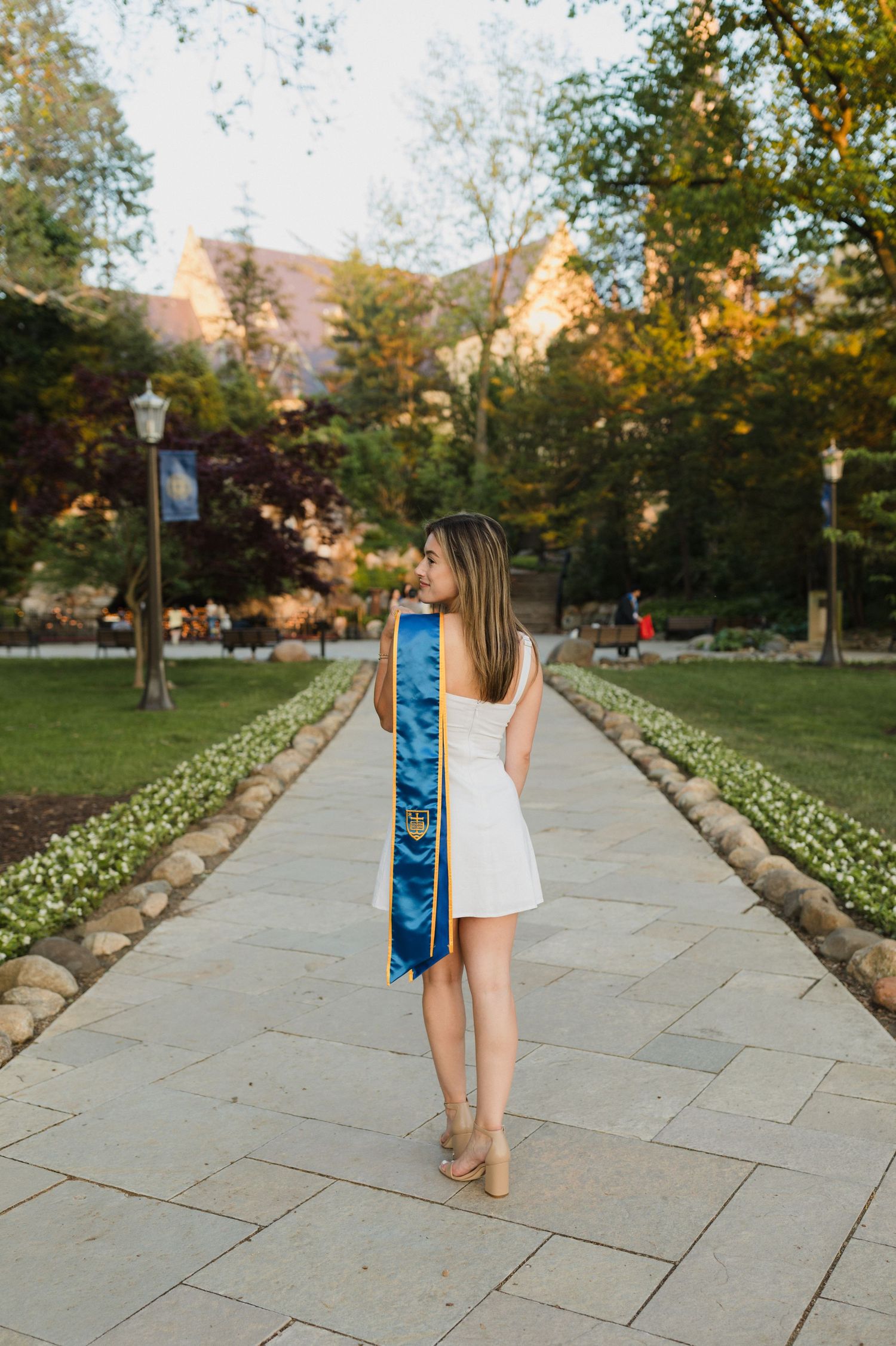 Graduate in white dress with blue graduation stole walking on stone path at sunset.