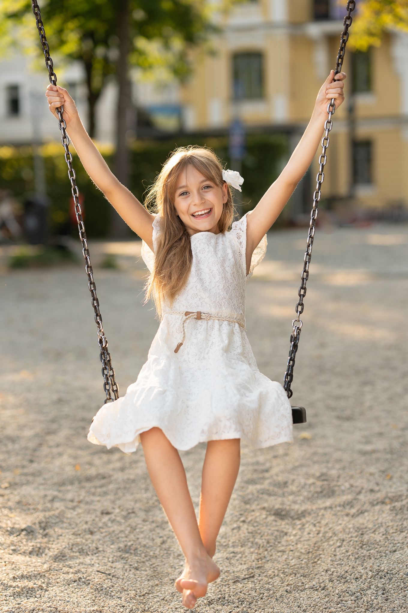 Kinderfotografie - Child in white dress joyfully swinging on playground swing with arms raised in summer.