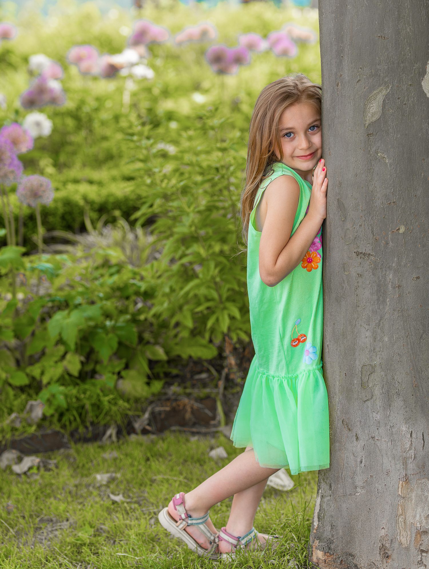 Kinderfotografie, Young girl in a green summer dress poses shyly against the trunk of a treer in a blooming garden.