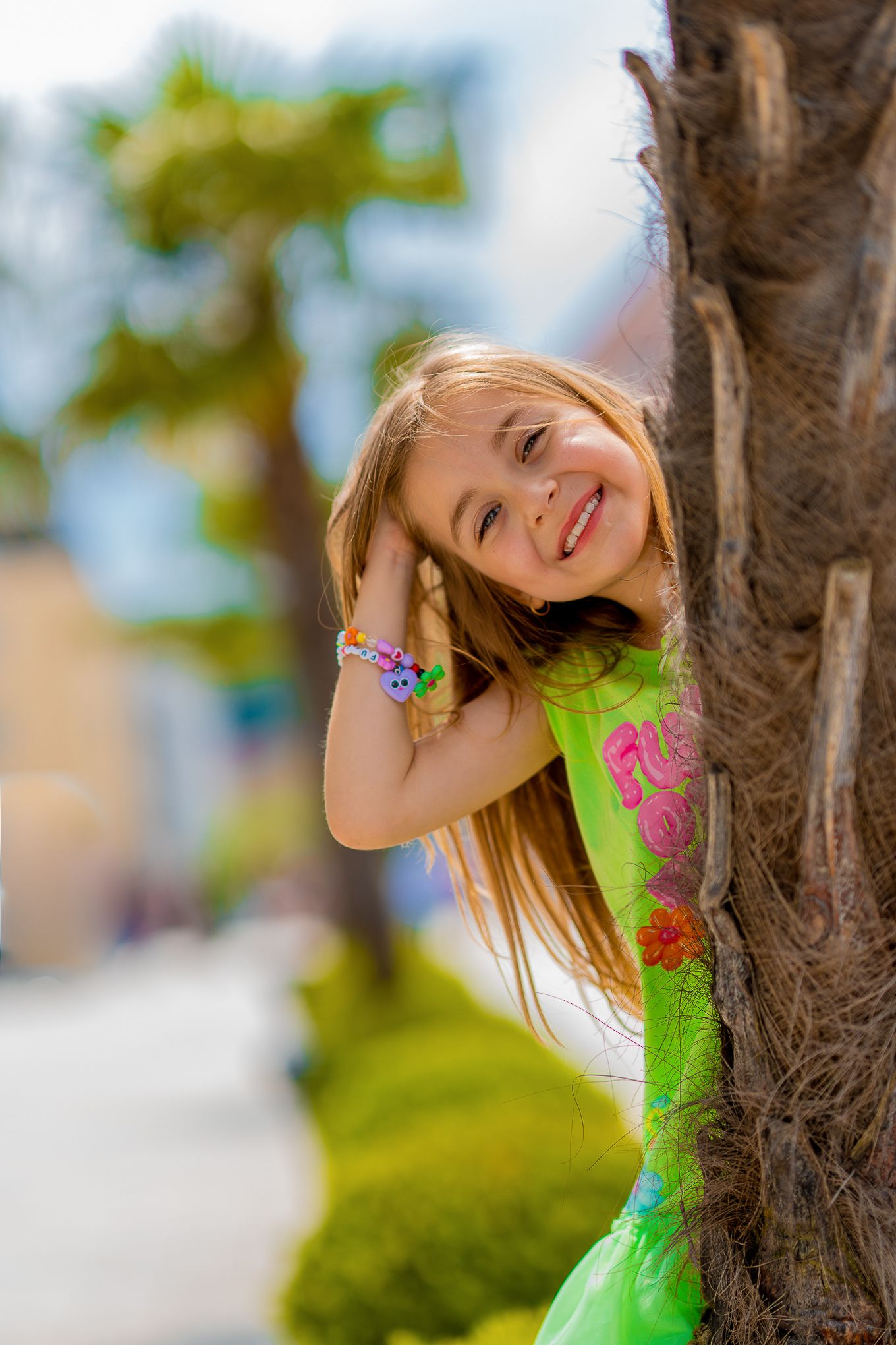 Kinderfotografie - Young girl in a bright green dress peeks playfully from behind a palm tree on a sunny day.