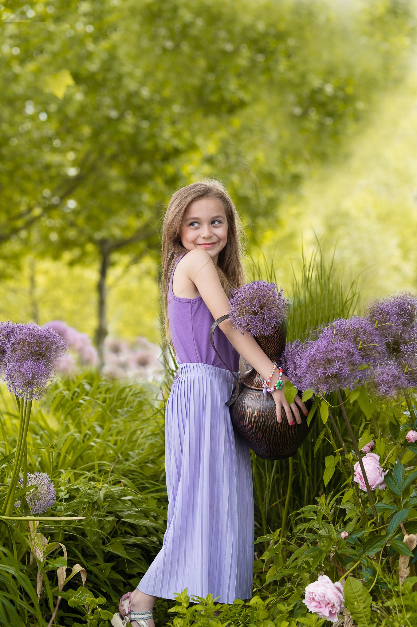 Kinderfotografie - Elegant garden scene of a girl with bronze vase and purple allium flowers in sunlit setting.