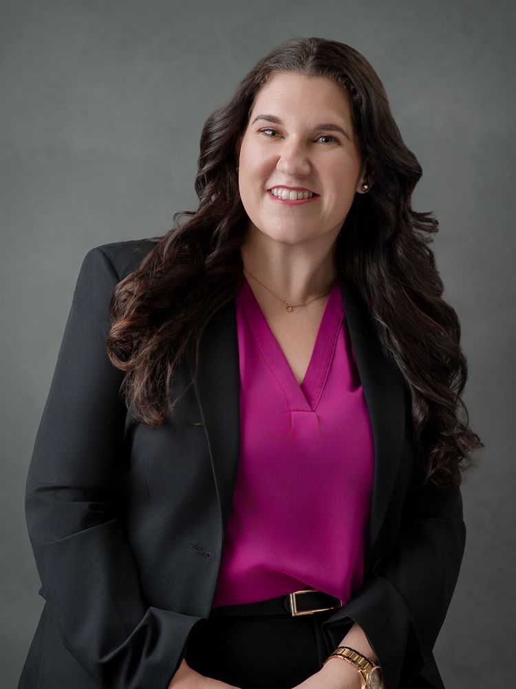 Professional woman in pink blouse and dark blazer smiling confidently against gray background.