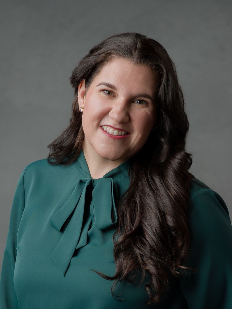 Professional headshot of a smiling woman with dark hair wearing a teal green bow-tie blouse against a gray background.