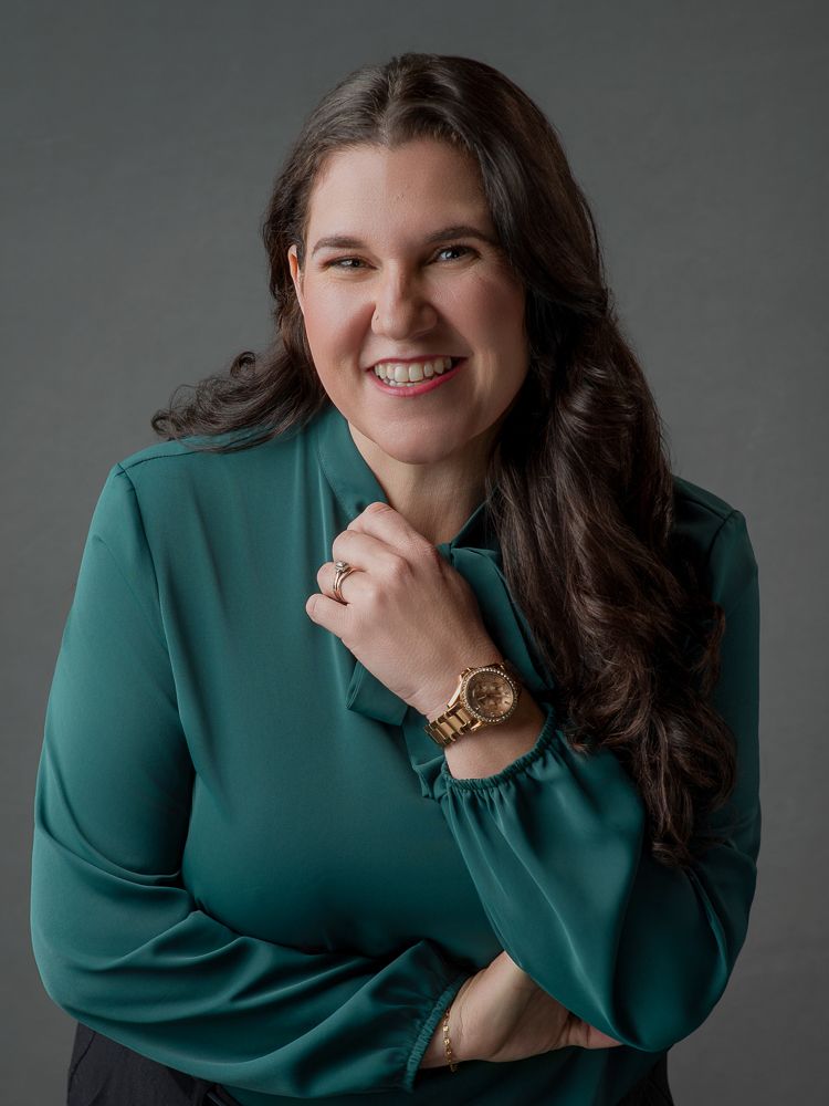 Professional woman in teal blouse smiling confidently, wearing a gold watch, posed against a gray studio background.
