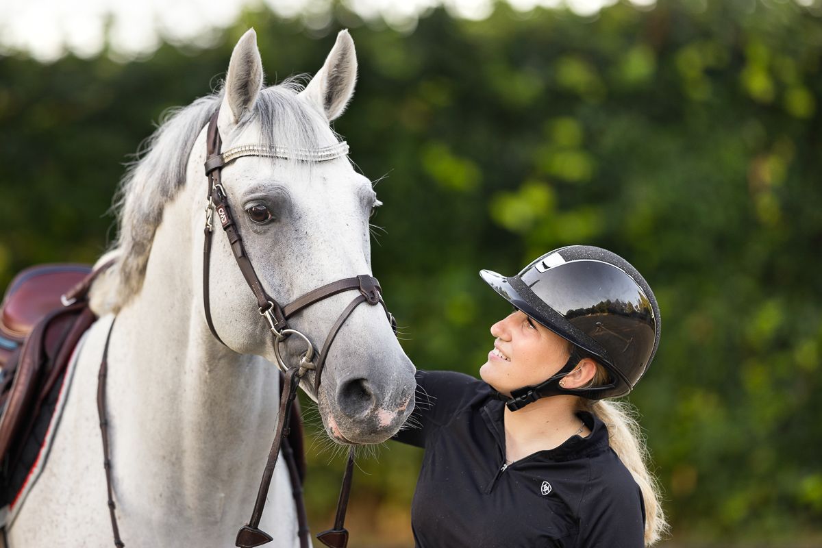 A rider in a black helmet looks up at their elegant white horse wearing a bridle in an outdoor setting.