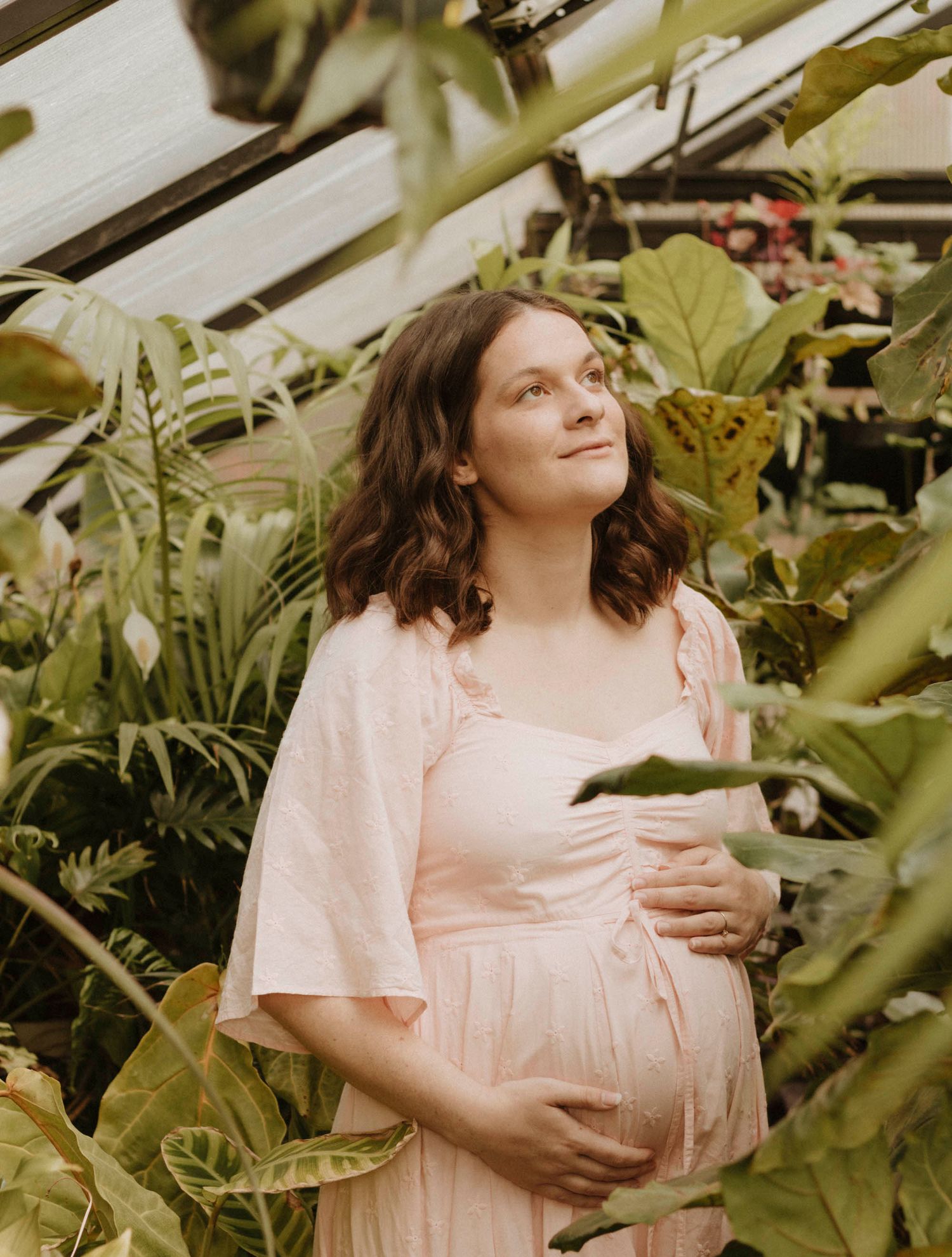 Greenhouse maternity photo among lush plants in pink flowy dress.