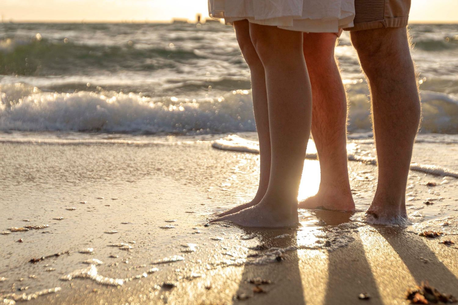 Feet standing in shallow ocean waves at sunset casting long shadows on wet sand.