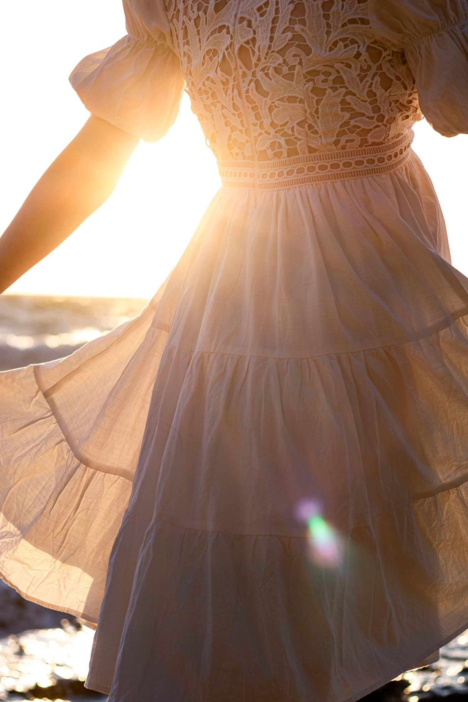 White lace dress twirling on beach at sunset with sun flare through fabric during a maternity shoot at Black Rock Beach