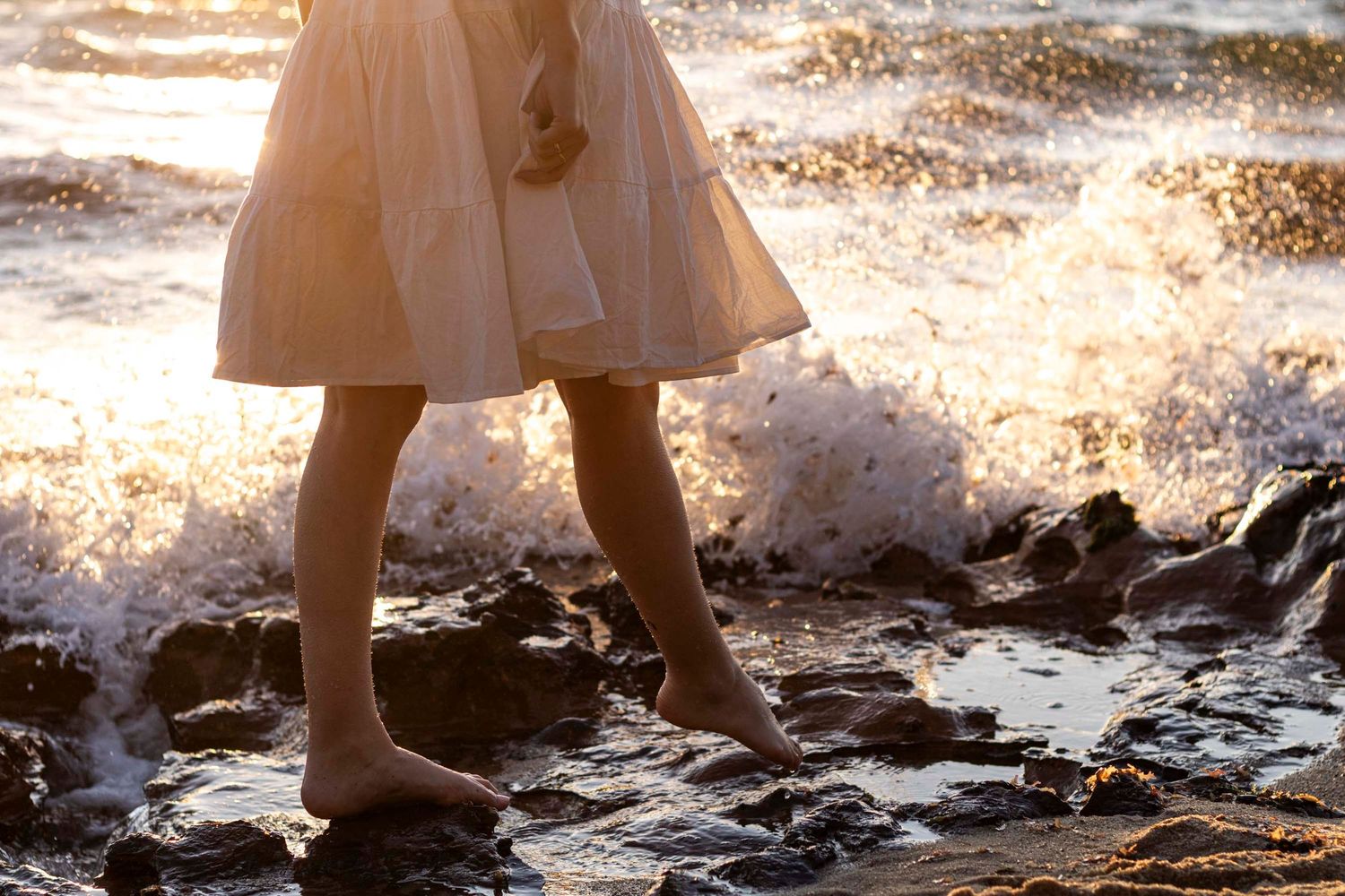 Bare feet in a flowing dress stand on rocky shore as ocean waves crash around them in golden sunlight.