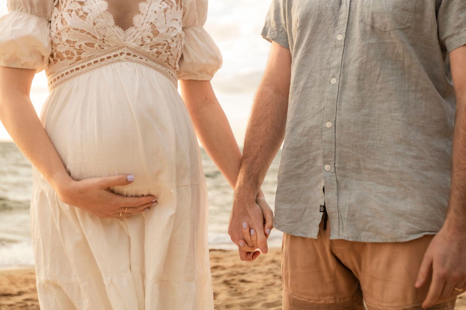 Close-up of expecting couple holding hands in flowing white dress and casual attire on beach.