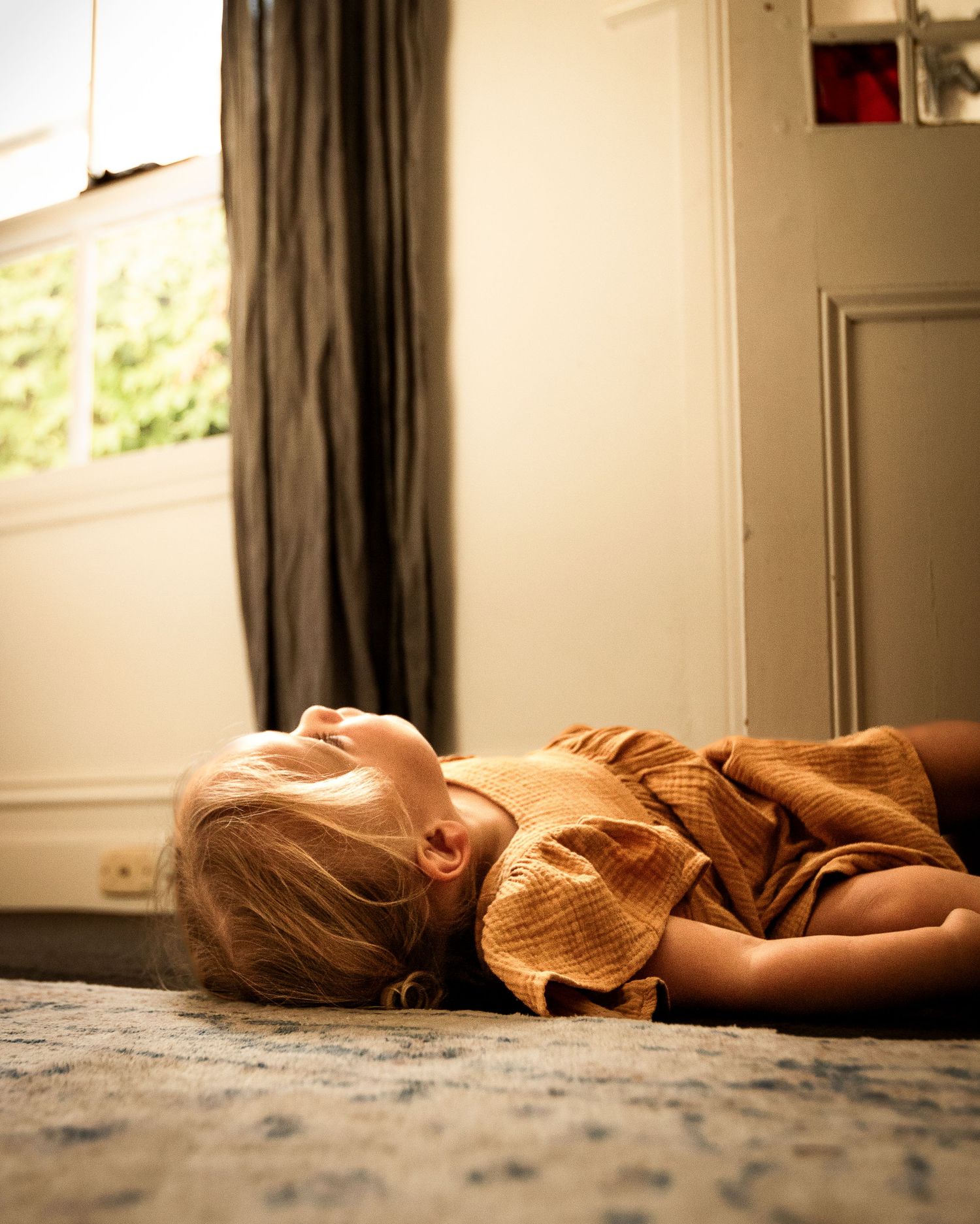 Child in orange clothing lying peacefully on carpet in sunlit room.