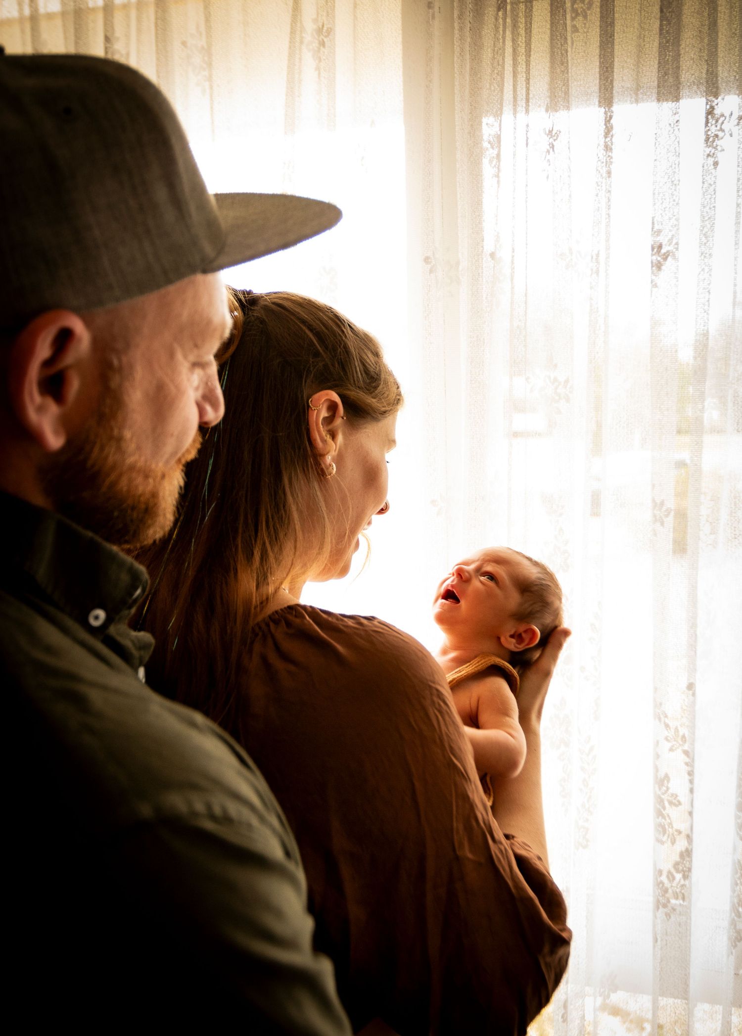 A tender family moment as parents stand with their newborn baby by a sunlit window.